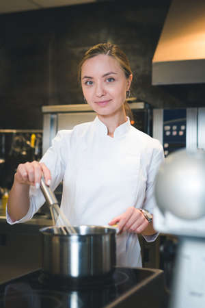 Portrait of confident and smiling young woman chef dressed in white uniform, professional kitchen are on background. restaurant kitchenの写真素材
