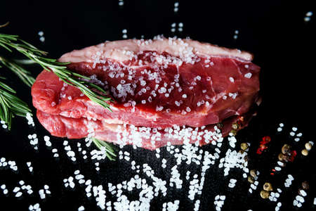 Raw organic beef meat with rosemary, seasonings, salt and red pepper on black background, close-up. Preparing to cook steakの写真素材