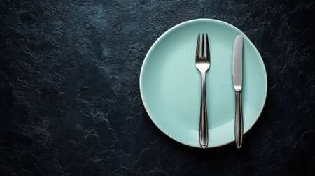 A minimalist composition showcasing a light blue plate with a fork and knife, set against a dark textured surface, ideal for culinary or dining themes.の素材