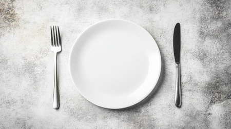 A minimalist table setting featuring a white plate, knife, and fork arranged on a textured concrete surface, perfect for showcasing culinary creations or dining concepts.の素材