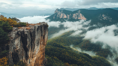 Wide view of a stone cliff jutting out above the forested mountain slopes with clouds drifting through the peaksの素材