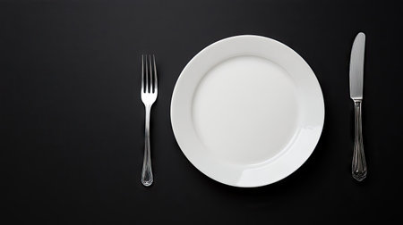 A white empty plate with a fork and knife neatly placed on a black background, creating a clean and minimalist dining setupの素材