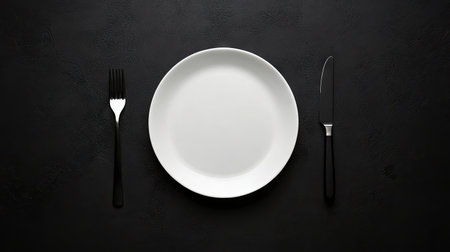 Minimalist table setup with a plate, knife, and fork on a black surface, ideal for food service or hospitality promotionsの素材