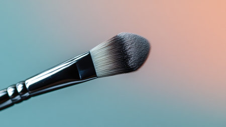 A close-up of a makeup brush tip with powder gently falling off the bristles, against a subtle gradient background.の素材