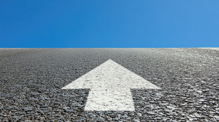 Long highway with clear white arrow painted on asphalt pointing forward under a bright blue skyの素材