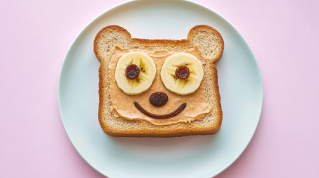 Fun animal face toast with peanut butter base, banana snout, and raisin eyes, served on a child-friendly pastel plate.の素材
