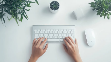 A tranquil overhead image showcasing hands typing on a sleek white keyboard, accompanied by a mouse and greenery, ideal for highlighting productivity and workspace aesthetics.の素材