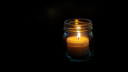 Single candle in a glass jar burning in the dark, soft glow illuminating the jar and creating a cozy ambiance.の素材