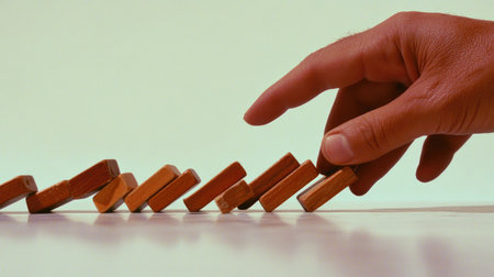 Close-up of a hand stopping a line of falling dominoes in mid-topple, capturing the moment of intervention and control on a light background.の素材
