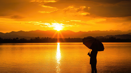 A striking silhouette of a person holding an umbrella as the sun sets behind them, casting a golden glow across a lake.の素材