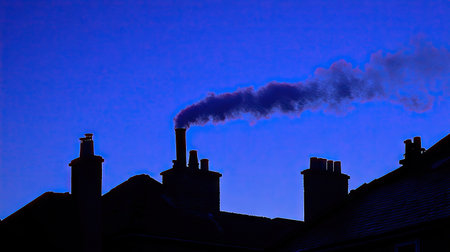 A chimney emitting a trail of white smoke in the early hours of the morning. The gentle smoke contrasts with the deep blue sky and silhouetted rooftops.の素材