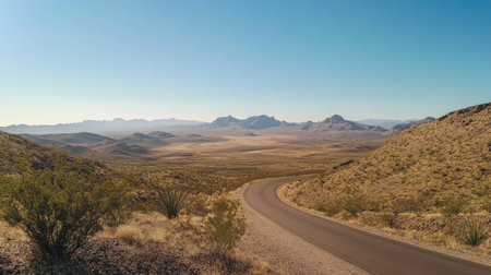 A wide shot of a desert road winding through the arid landscape, with distant mountains and a clear blue sky, inviting adventure.の素材