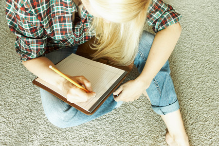 A blonde woman in a checkered red-green shirt and jeans sits on the floor on the carpet and writes in a folder with a pencilの写真素材