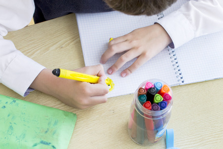 A boy, a schoolkid in white shirt is drawing with yellow marker in a notebook . Many colorful markers are on the table.の写真素材