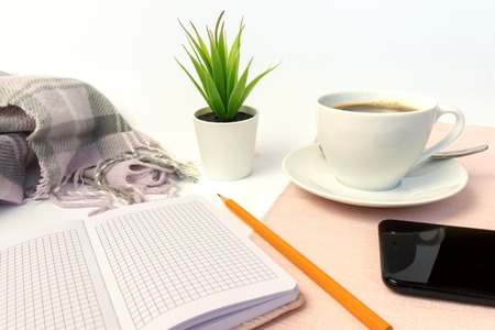 White ceramic cup of coffee, notebook, pencil, green plant and smartphone on white table, covered...