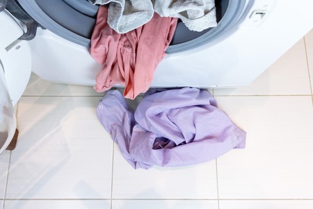 Washing machine full of dirty laundry and dirty clothes on a bathroom floor.