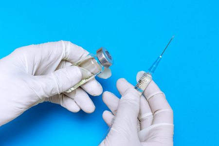 Hands of a nurse in white gloves holding syringe and transparent vial on blue background.