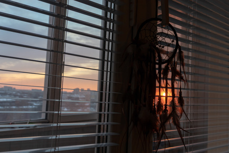 Handmade dreamcatcher hanging by the window with blinds in sunset twilight. Black silhouette of...