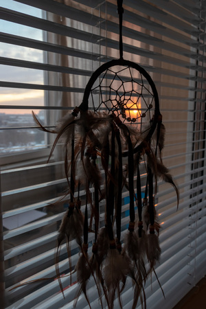 Handmade dreamcatcher hanging by the window with blinds in sunset twilight. Black silhouette of traditional magic amulet for dream protection and bad spirits.の写真素材