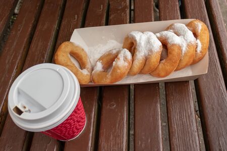 Closeup of beignet pastries, donuts in a paper takeaway box and a cup of coffee on a bench in park....