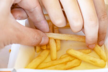 Hands of a hungry people taking out fried potatoes out of a take away out paper box and eating it on the summer terrace of the city fast food restaurant, close up view.の写真素材