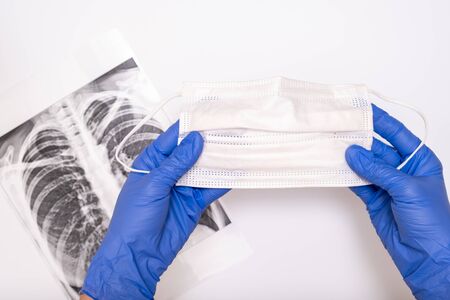 A doctor in medical gloves holding a protactive face surgical mask, an x-ray of lungs on backdrop,...