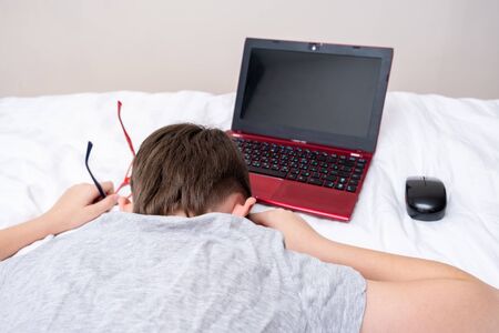 Bored and tired boy lying on a bed with a laptop after doing his homework, distant remote education during quarantine.の写真素材
