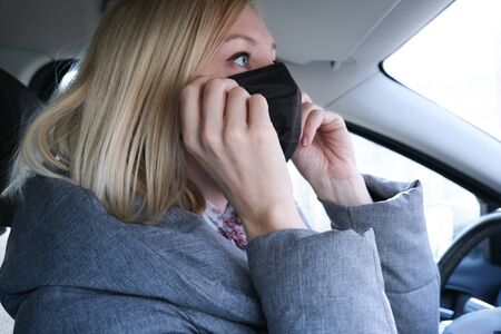 Caucasian european woman sitting in a car and putting on black surgical medical face mask as a way...