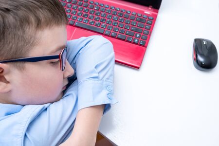 A tired boy, schoolboy in blue shirt sitting by the table with a laptop on it at home, difficulties...