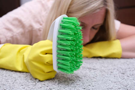 Woman in yellow gloves with a green brush is so tired of cleaning and brushing carpet, removing stains and wool from it and doing routine homework, that she lay down on the floor and sleepの写真素材
