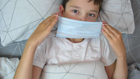 A portrait of a sick caucasian boy lying in bed and putting on a medical protective mask, illness and flu concept.の写真素材