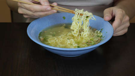 Woman sitting by the table and eating instant egg noodles with chopsticks from a ceramic dish, quick...