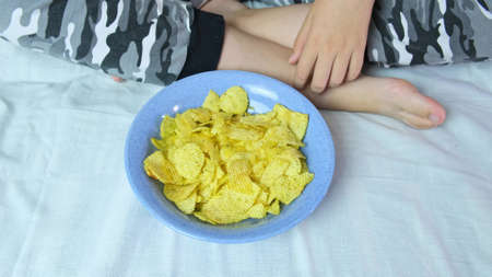 A boy sitting on a sofa at home and eating crispy potato chips from a bowl, close up view.