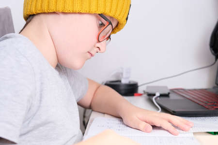 A boy in a trendy yellow knitted hat sitting by the table at home and doing homework, he using a laptop and whiting in a notebook.の写真素材