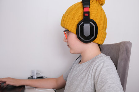 A gamer boy in a trendy knitted cap and heaphones sitting by the table and playing multiplayer computer game on laptopの写真素材