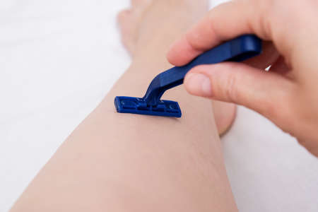 Woman shaving legs with a blue disposable plastic razor close up, unwanted hair removing