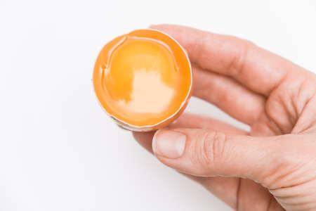 A hand holding brown chicken egg with yellow yolk is half broken against white background