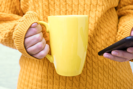 Unrecognizable woman in yellow autumn knitted sweater holding a big cup, mug of coffee and using smartphone, fall morning relaxation concept.の写真素材