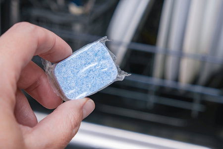 A hand holding blue dishwasher soap tablet in a water-soluble packaging on a dishashind machine...