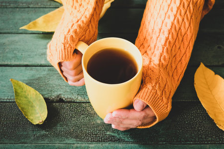 Female woman hands wearing yellow knitted sweater pullover holding a mug of coffee, hot beverage agaunst green wooden background with autumn leavesの写真素材