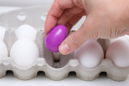A person is carefully picking up a vibrant purple egg from a carton filled with several white eggs of varying sizes and shapesの写真素材