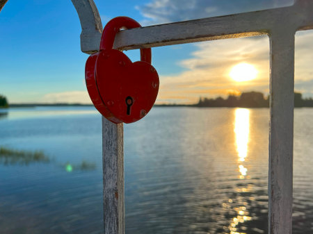 A vibrant red heartshaped padlock has been securely attached to a sturdy railing that overlooks a picturesque lake settingの写真素材