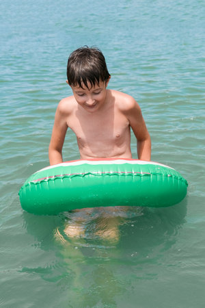 A joyful boy plays in blue water with a green float, embodying summertime happiness and carefree funの写真素材