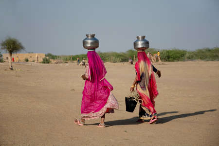 Two woman carrying water in steel vessels on head  in desert, Khuri, Rajasthan, Indiaのeditorial素材