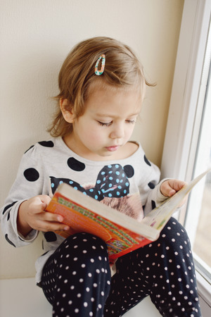 little girl is reading a book near the windowの写真素材