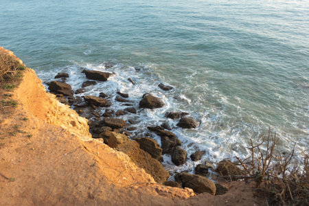 Ocean waves crashing on rocks below sandy cliff at sunsetの写真素材