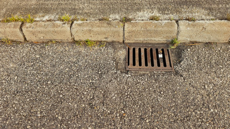Close-up of a sewer grate draining water on an asphalt road next to a concrete curbの写真素材