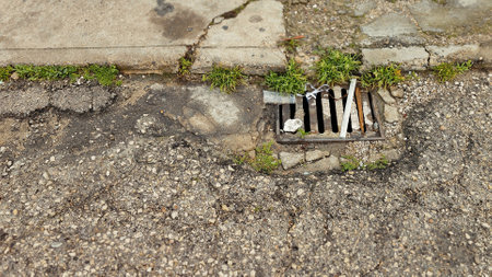 Damaged asphalt road with a sewer grate, weeds, and debris, showing urban decay and neglectの写真素材