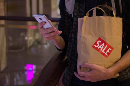 Man hold brown paper bag with SALE sticker on it in one hand and use smartphone with another Close up HDの写真素材