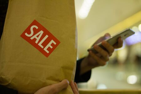 Man hold brown paper bag with SALE sticker on it in one hand and use smartphone with another Close up HDの写真素材